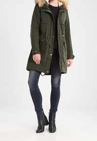 Olivově zelená parka s umělou kožešinou na kapuci, se zapínáním na zip a patentky, dvěma předními kapsami a stahovacími šňůrkami v pase, s volnějším střihem.