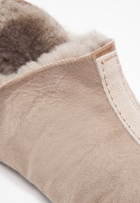 Beige semsket tøffel med et mykt, fôr av pels innvendig, en avrundet form, og en sydd detalj langs åpningen. Merket synlig.