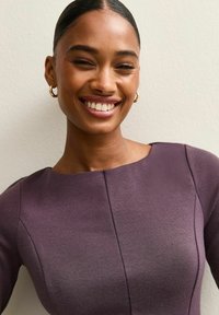 Mujer sonriendo con el cabello recogido y liso, que lleva pendientes de aro dorados y una blusa texturizada morada, posa frente a un fondo claro y liso.
