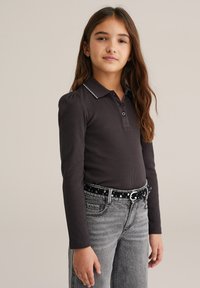 Tricou polo cu mânecă lungă, de culoare gri închis, cu o textură ribbă, având o cusătură contrastantă albă la guler. Asortat cu blugi gri și o curea cu puncte.