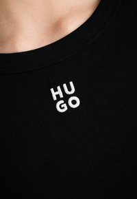 Gros plan d'un t-shirt noir côtelé à col rond avec des lettres blanches brodées "HU GO" près de la zone de la clavicule.