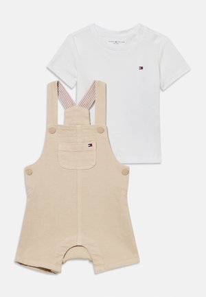 Λευκό κοντομάνικο μπλουζάκι για νήπια με μικρό λογότυπο και μπεζ σορτς ολόσωμο με ρυθμιζόμενες τιράντες και εμπρόσθια τσέπη, και τα δύο με σήμανση.