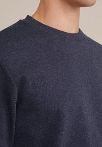 Mörkblå texturerad tröja med rundad halsringning och ventilerande tyg. Färgbild av materialet som visar subtila stickmönster.