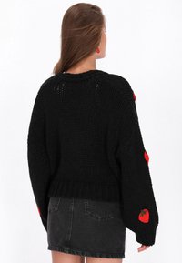 Kvinna som vänder bort ansiktet, iklädd en svart grovstickad tröja med röda jordgubbsmönster på ärmarna och en urtvättad svart denimkjol.