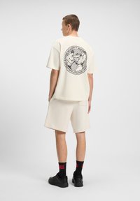 Cremefarvet oversized T-shirt med grafisk print på ryggen. Sammen med matchende cremefarvede shorts, sorte sokker og sorte sneakers.