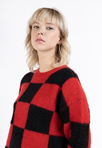 Pulover tricotat în carouri roșii și negre, cu guler rotund și țesătură texturată. Model masiv cu culori contrastante.