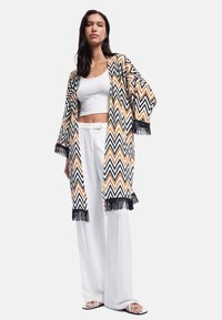 Kvinde iført en hvid crop top, brede hvide bukser, åben cardigan med zigzag mønster og sort frynser samt hvide sandaler, stående på hvid baggrund.