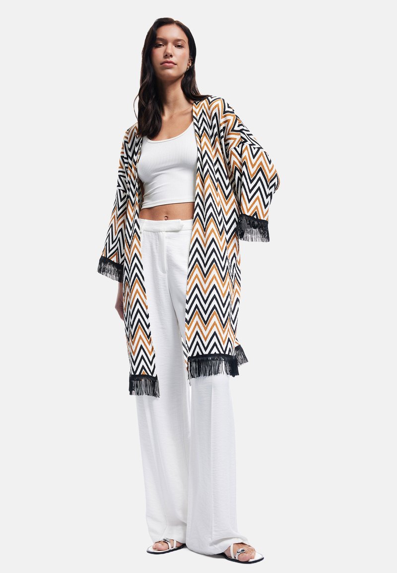 Kvinde iført en hvid crop top, brede hvide bukser, åben cardigan med zigzag mønster og sort frynser samt hvide sandaler, stående på hvid baggrund.