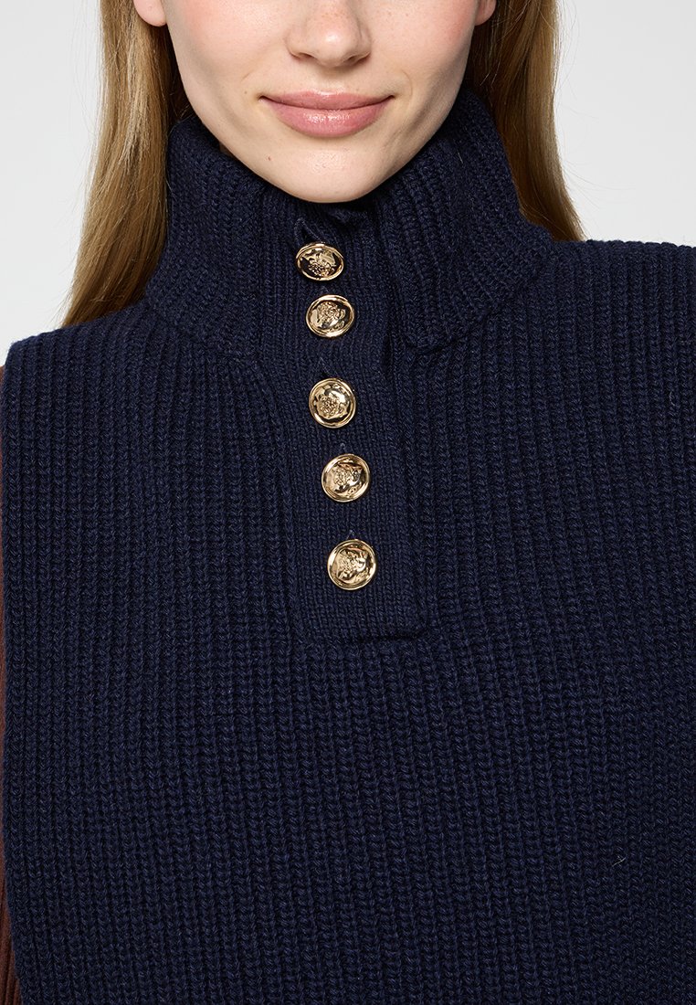 Pull-over en tricot marine avec un col haut et cinq boutons décoratifs dorés au niveau du cou. Tissu texturé avec un motif côtelé.