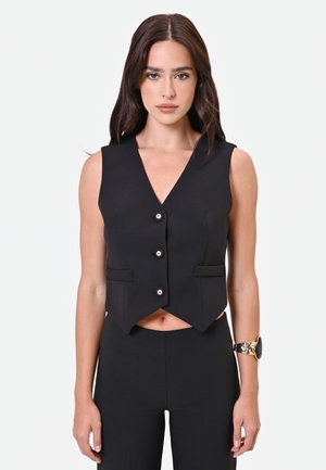 Donna con capelli castani lisci indossa un gilet nero senza maniche con bottoni e pantaloni abbinati, in piedi davanti a uno sfondo bianco semplice.