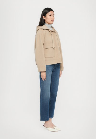 Veste à capuche beige avec une poche avant, associée à un jean bleu droit. Le mannequin porte des chaussures à enfiler crème. Design simple et structuré.