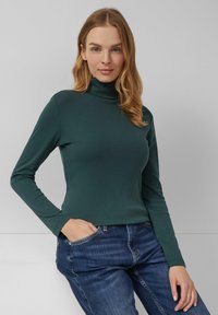 Donkergroene coltrui van zachte stof, met lange mouwen en een aansluitend ontwerp, gecombineerd met blauwe jeans.