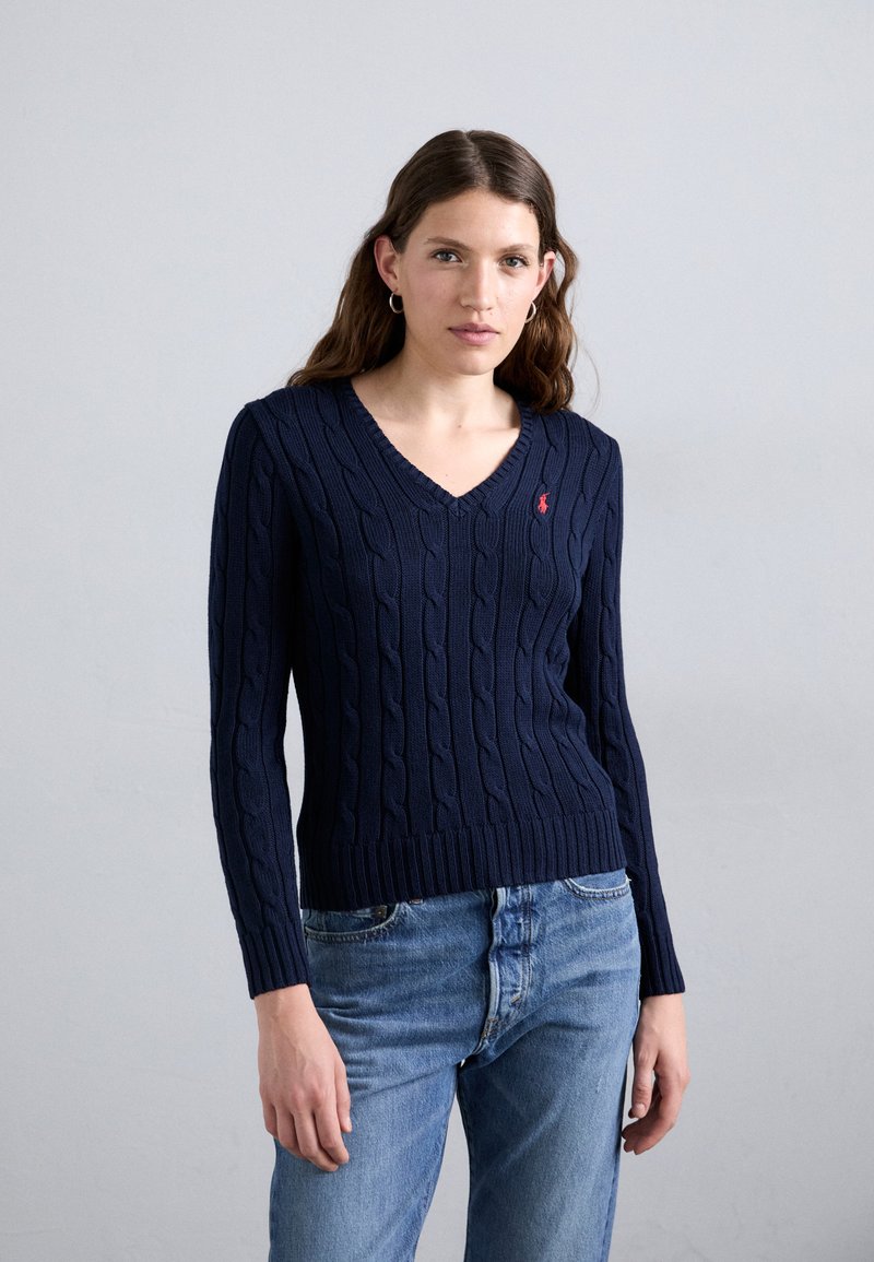 Navy cable-knit sweater with a V-neck and ribbed hem, featuring a small red logo on the chest and long sleeves. Paired with blue denim jeans.