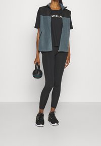 Conjunto deportivo que incluye un chaleco gris con cremallera sobre una camiseta negra de manga corta, leggings negros y zapatillas de deporte negras. Sosteniendo una kettlebell.