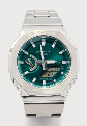 G-Shock Uhr mit einem Edelstahlarmband, türkisfarbenem Zifferblatt, silbernen Akzenten und digitaler Anzeige. Die Funktionen sind um das Ziffernblatt herum beschriftet.