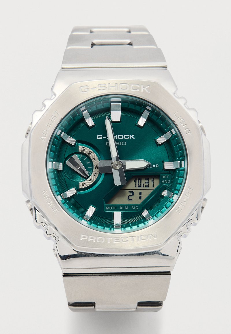 G-Shock kell, millel on roostevabast terasest rihm, olnud tealik nägu, hõbedased aktsendid ja digitaalne ekraan. Funktsioonid on tähistatud diali ümber.
