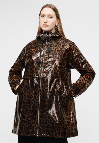 Νεαρή γυναίκα με ίσια μαλλιά, φορώντας μια γυαλιστερή αδιάβροχη καμπαρντίνα με κουκούλα και λεοπάρ σχέδιο, με τα χέρια στις τσέπες, πάνω σε ένα απλό φόντο.
