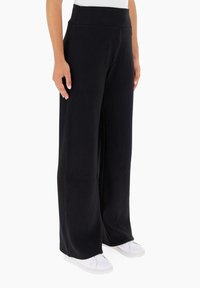 Pantaloni neri a coste con gamba larga e alta vita, realizzati in un materiale morbido, abbinati a sneakers bianche. Presentano una texture verticale.