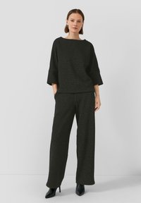 Femme debout portant un haut noir texturé ample avec des manches larges, un pantalon large assorti et des bottines à talons aiguilles noires.