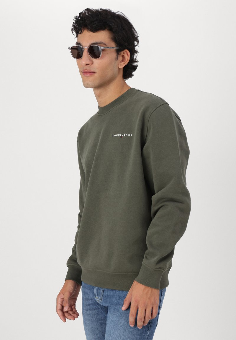 Felpa verde oliva con scollatura a girocollo e polsini a costine, con un piccolo logo bianco. Indossata con jeans blu e occhiali da sole neri.