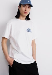 Hvit bomulls t-skjorte med kort erme, med et blått grafisk logo på venstre bryst, parret med svarte shorts og en blå caps.