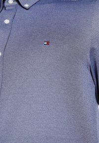 Camicia blu intrecciata con colletto abbottonato, presenta un piccolo logo Tommy Hilfiger in rosso, bianco e blu sul petto. Tessuto texturizzato.