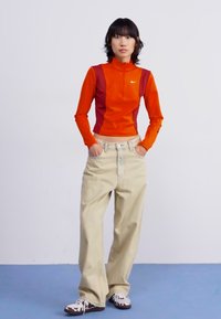 Långärmad orange och vinröd träningströja med hög hals, matchad med beigefärgade baggy byxor och mönstrade sneakers. Enkel, modern design.