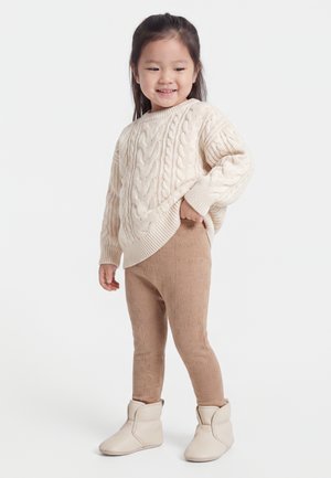 Ung pige smiler, iført en cremefarvet kabelstrikket sweater, lysebrune mønstrede leggings og lysebeige bløde støvler, står mod en ensfarvet baggrund.