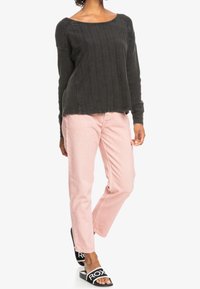 Persoon in een donkergrijze trui met lange mouwen, lichtroze jeans en zwarte instapsandalen met het witte "ROXY"-logo op een witte achtergrond.