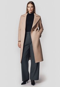 Beżowy wełniany płaszcz z wyłogowym kołnierzem, podwójnym zapięciem i bocznymi kieszeniami, noszony na czarnym golfie i szarych szerokich spodniach.