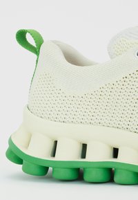 Sapatilha branca em malha com um upper texturizado, detalhes em verde, e uma sola acolchoada com cápsulas verdes arredondadas para absorção de choques.