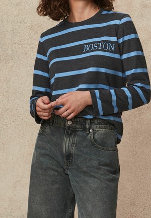 Šedá košile s dlouhými rukávy a modrými horizontálními pruhy, se výšivkou "BOSTON", v kombinaci s džínami s vysokým pasem v tmavé barvě.