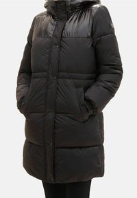 Czarna, długa puchowa kurtka z błyszczącymi panelami na ramionach, z przodu na zatrzaski, z kieszeniami po bokach i wysokim kołnierzem noszona przez osobę.