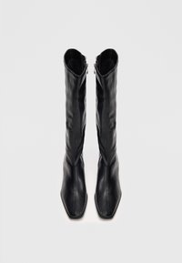 Une paire de bottes en cuir noires hautes avec des orteils carrés et des fermetures éclair sur les côtés, vues du dessus sur un fond clair.