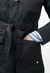 Main dans la poche d'un manteau noir ceinturé, portant un chemisier à manches longues bleu et blanc rayé en dessous.