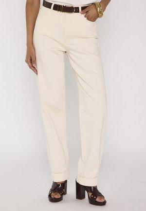 Personne portant un pantalon droit crème taille haute, retroussé aux chevilles, ceinture en cuir marron et talons compensés marron foncé à bouts ouverts.