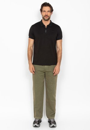 Mężczyzna stojący przodem, ubrany w czarną koszulkę polo z krótkim rękawem, oliwkowozielone spodnie oraz czarne i szare buty sportowe.