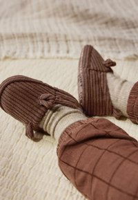 Des chaussons en velours côtelé marron présentent une texture douce, avec des accents noués à l'avant. Portés avec des chaussettes beige clair et un pantalon assorti marron à motifs.