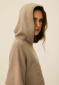 Beżowy, dziany hoodie z dużym, miękkim kapturem. Posiada prążkowane mankiety i dół oraz wytłaczane detale z przodu. Gładka tekstura.