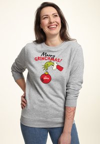 Sweatshirt gris avec un design de cartoon et le texte "Joyeux Grinchmas !", représentant le Grinch tenant une ornament rouge. Texture douce, coupe décontractée.