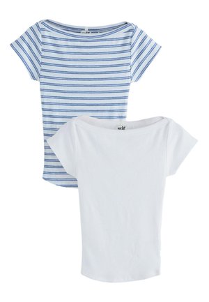 Frau trägt ein enganliegendes weißes Kurzarmhemd und hellblaue Shorts mit Kordelzug, Hände in den Taschen, vor einfachem Hintergrund.