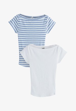 Kvinde iført en tætsiddende hvid kortærmet skjorte og lyseblå shorts med snor og hænderne i lommerne mod en ensfarvet baggrund.