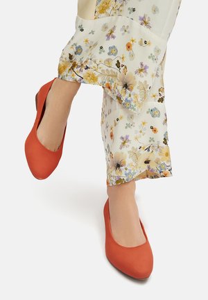 Nohy v květinových širokých kalhotách s jasně oranžovými nízkými podpatky na bílém pozadí.