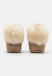Bruna mocka tofflor med en fluffig krämfärgad pälskant. Mjuk textur, rundad form och synliga sömnadsdetaljer vid hälen.