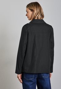 Chaqueta negra con cuello, costuras visibles y detalles de botones en los hombros. La tela parece suave. Lleva puesta sobre jeans azul oscuro.