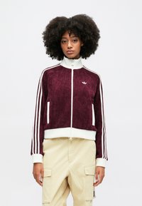 Burgundijas krāsas zip-up jakas ar baltām svītrām, ribota apkakle un piedurknes, mīksta tekstūra un divas priekšējās kabatas. Mazs logs krūšu daļā.