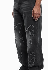 Μαύρο τζιν από denim με φθαρμένα στοιχεία, που περιλαμβάνουν σχισμένα τμήματα και ξεφτισμένα άκρα. Χαλαρή εφαρμογή και ελαφριά ξεθώριαση σε όλο το ύφασμα.