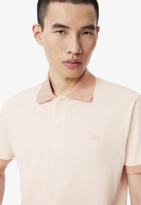 Paita polo berwarna merah muda muda dengan kerah peach yang kontras. Memiliki kain bertekstur, placket dua tombol, dan logo bordir di dada.