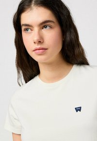 Camiseta de algodón crema con cuello redondo, mangas cortas y un pequeño logo bordado azul en el pecho. Tejido suave y ligero.
