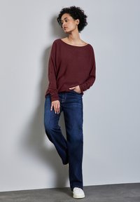 Bordeaux langemouwtop met wijde halslijn, gecombineerd met donkerblauwe spijkerbroeken en witte sneakers. Model poseert tegen een lichtgrijze muur.